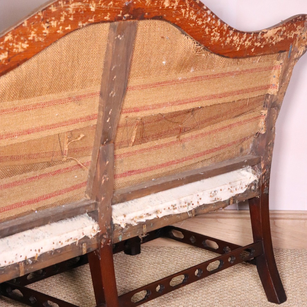 An antique Chippendale style mahogany settee featuring a serpentine camel back, scrolled arms, pierced stretchers, and exposed base lining fabric. View X