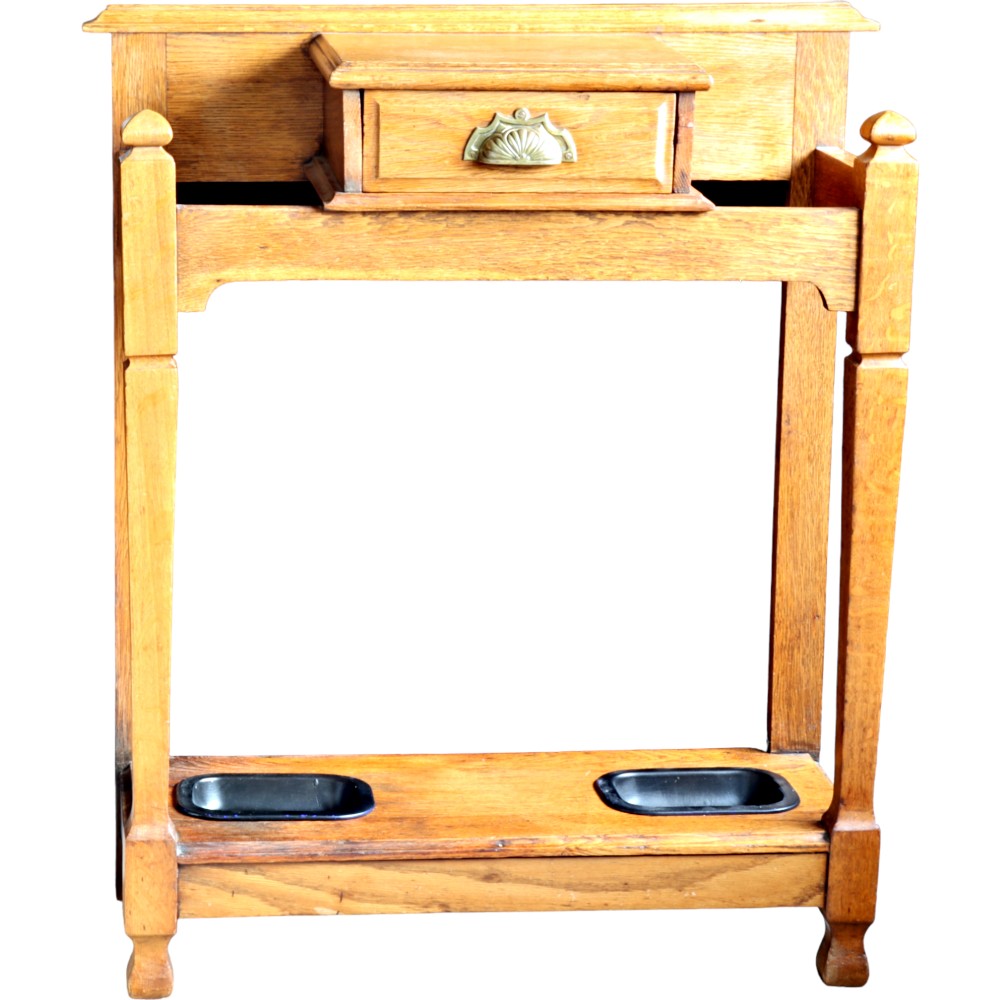 An antique oak hall stick stand featuring a central glove drawer with a brass cup handle, upper rails, square posts with finials, and a lower shelf fitted with two black drip trays.