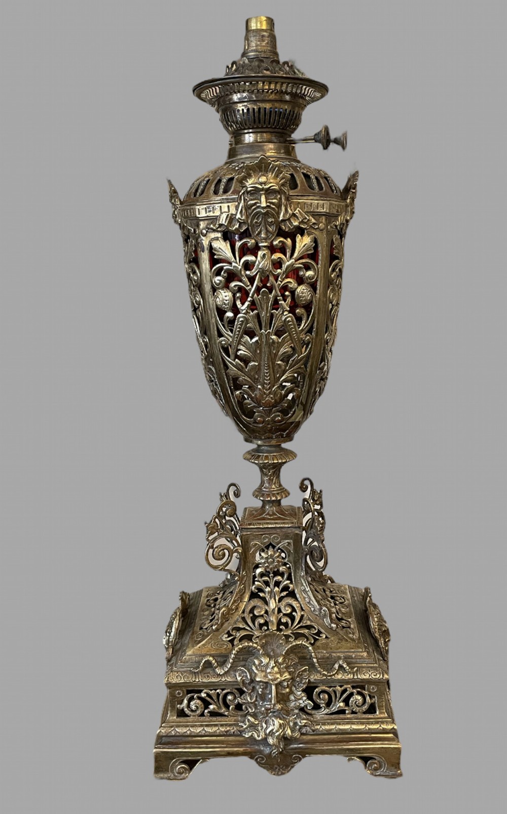 An ornate antique brass oil lamp featuring an intricate pierced metal reservoir holder decorated with mask and leaf motifs over a square stepped base.