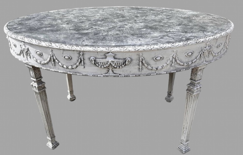 A late 19th-century oval centre table featuring a distressed grey painted finish, carved urn and swag apron details, and fluted tapered legs.