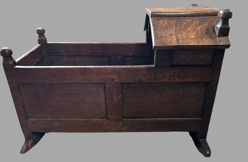 An early 18th-century dark oak rocking cradle featuring panelled sides, a hooded head section, turned corner finials, and solid wooden rockers.