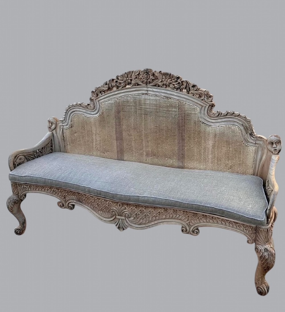 An Anglo-Indian carved bleached wooden settee featuring a woven cane backrest, decorative cresting, carved terminal head motifs, scroll legs, and a fitted grey linen cushion.