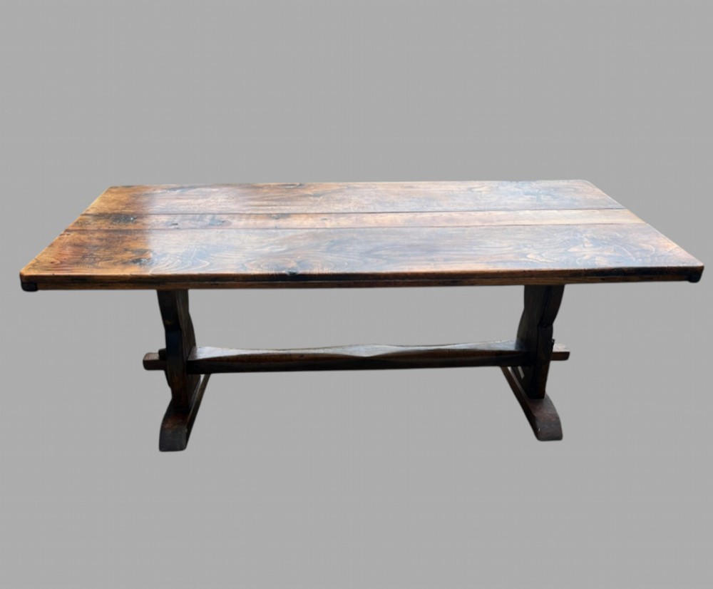 A rectangular wooden refectory dining table featuring a plank top and trestle base with a central stretcher in warm brown tones.