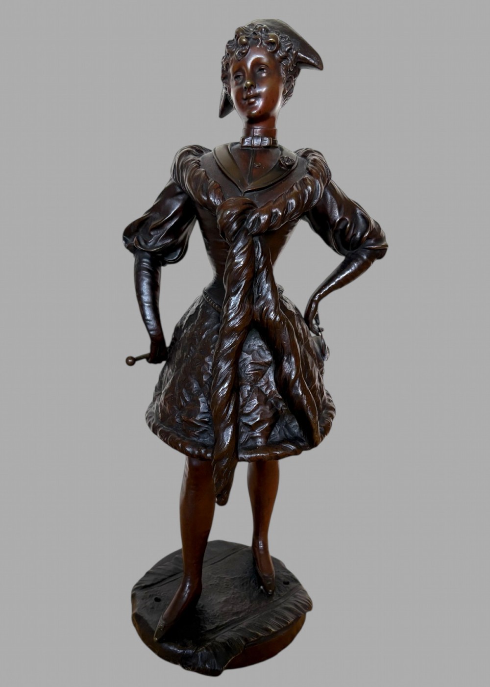 A dark patinated bronze figurine depicting a young woman standing in early 20th-century costume with a fur scarf and hat on an integral base.
