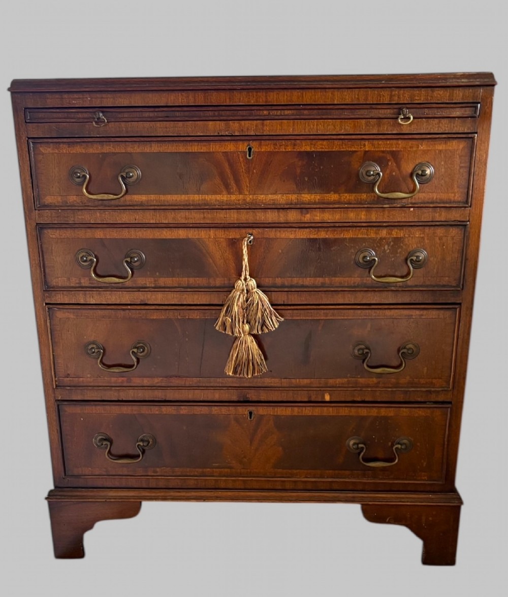 A front view of a compact antique mahogany bachelors chest featuring multiple graduated drawers, brass drop handles, and bracket feet.