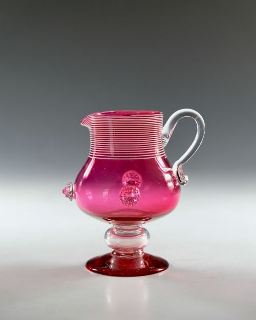 fig 1. A Victorian cranberry glass cream jug with white threaded details on the neck, a clear applied handle, glass prunt decorations, and a pedestal base.