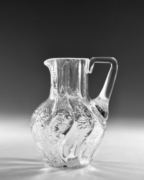fig 1. An antique clear glass jug featuring a bulbous swirl-moulded lower body with wheel-engraved floral motifs and a fitted handle.