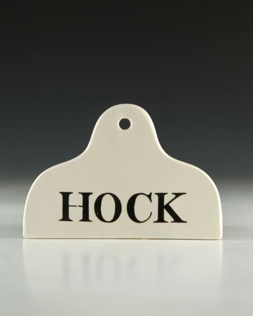 fig 1. A cream glazed ceramic wine bin label with an arched top, single hanging hole, and black printed serif text reading HOCK.