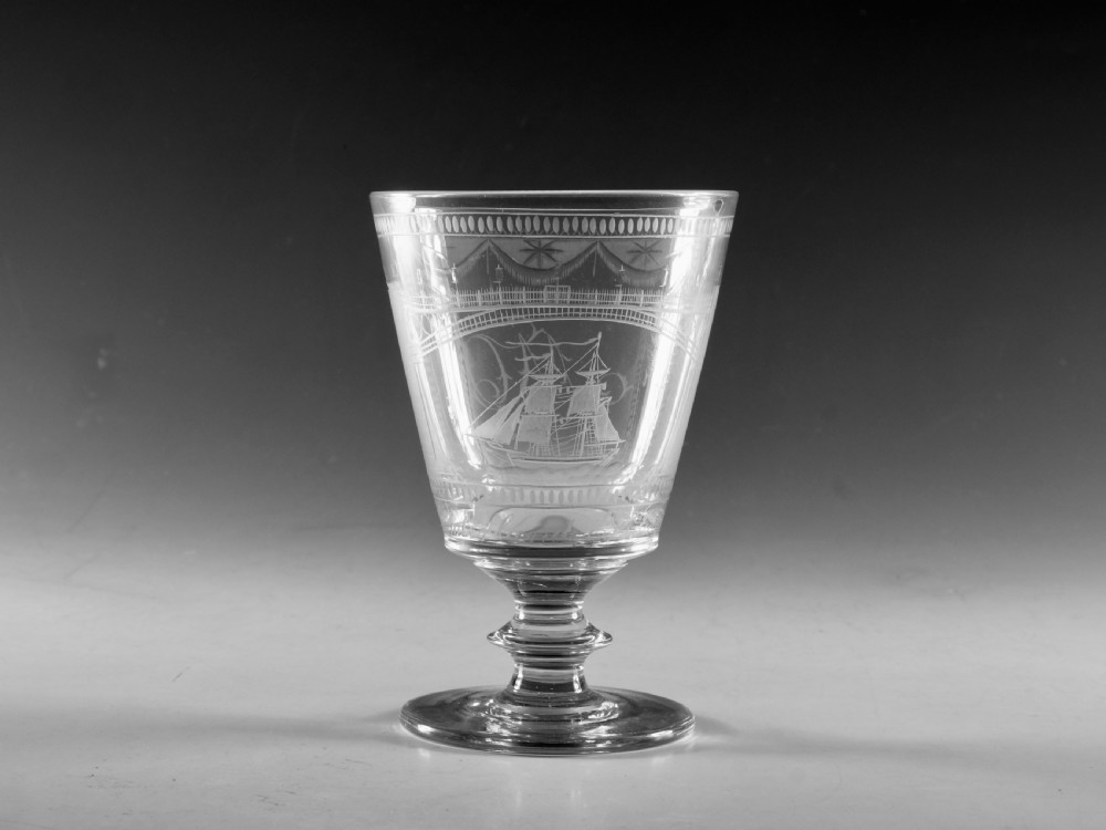 An antique clear lead glass rummer featuring detailed wheel engraving of a sailing ship beneath a bridge, set on a knopped stem and circular foot.