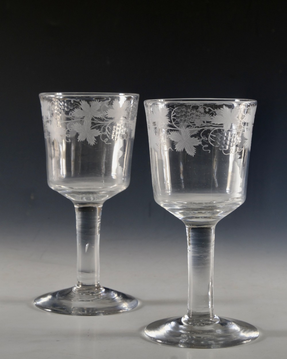 A pair of 18th-century clear lead glass goblets featuring plain straight stems and wheel-engraved grapevine motifs around the rims. View VI