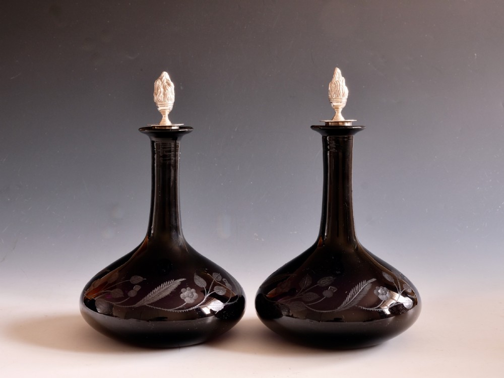 A pair of dark amethyst glass decanters featuring wheel-engraved botanical motifs and topped with silver flame-shaped stoppers.