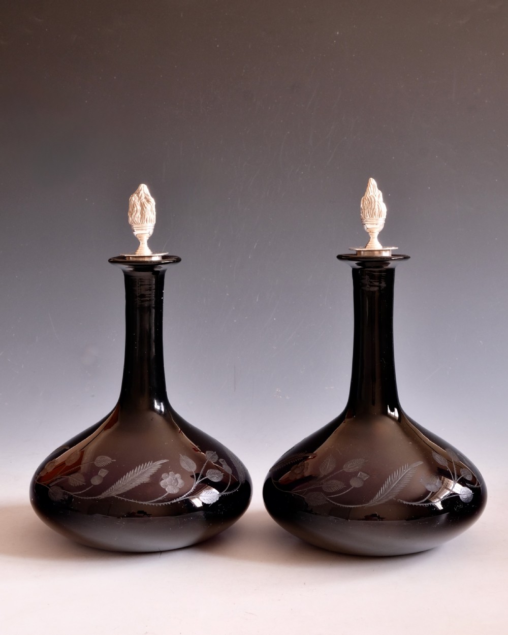 A pair of dark amethyst glass decanters featuring wheel-engraved botanical motifs and topped with silver flame-shaped stoppers. View II