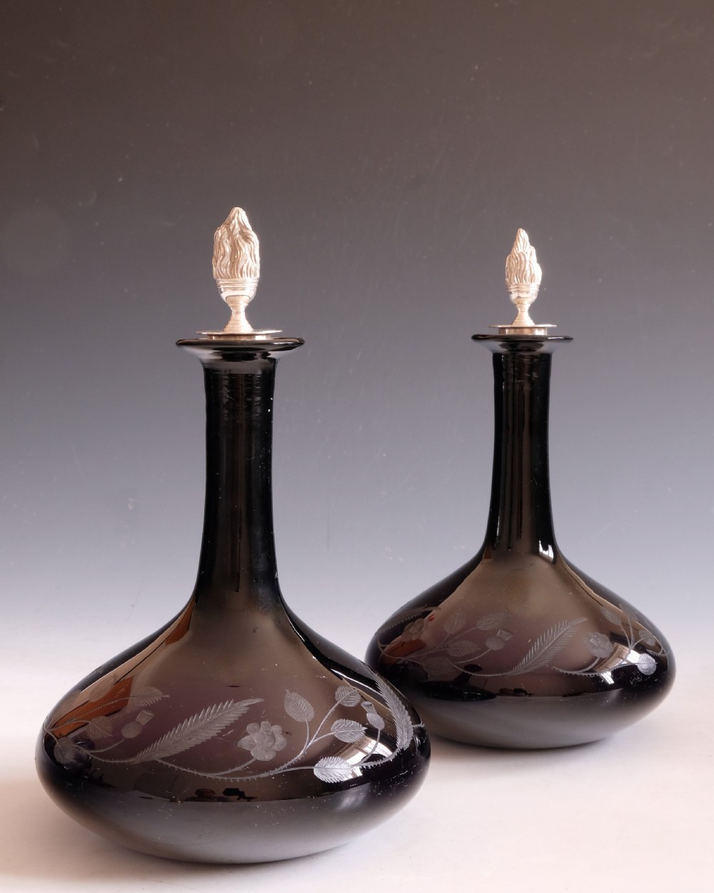 A pair of dark amethyst glass decanters featuring wheel-engraved botanical motifs and topped with silver flame-shaped stoppers. View IV