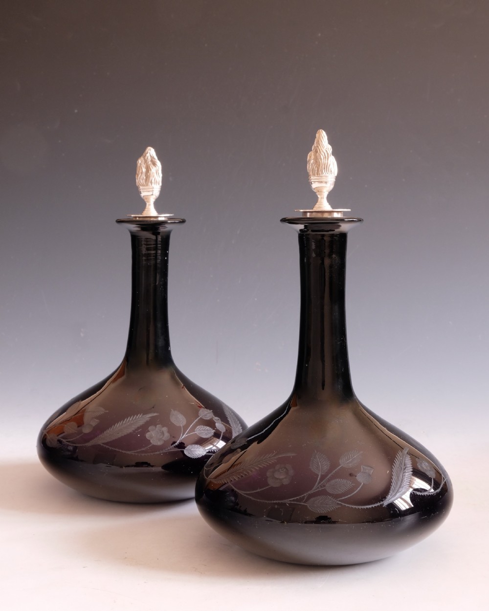 A pair of dark amethyst glass decanters featuring wheel-engraved botanical motifs and topped with silver flame-shaped stoppers. View V