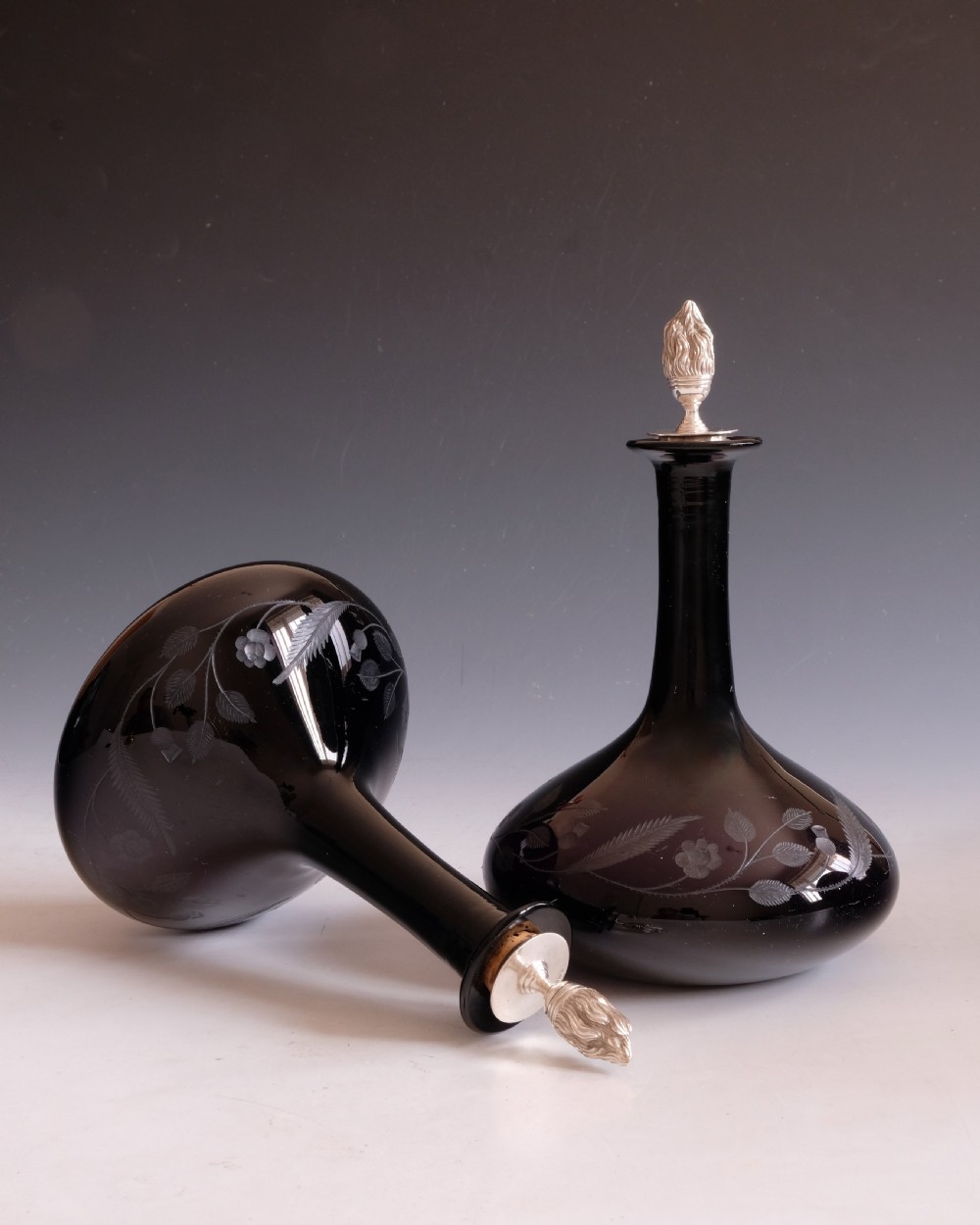 A pair of dark amethyst glass decanters featuring wheel-engraved botanical motifs and topped with silver flame-shaped stoppers. View VII
