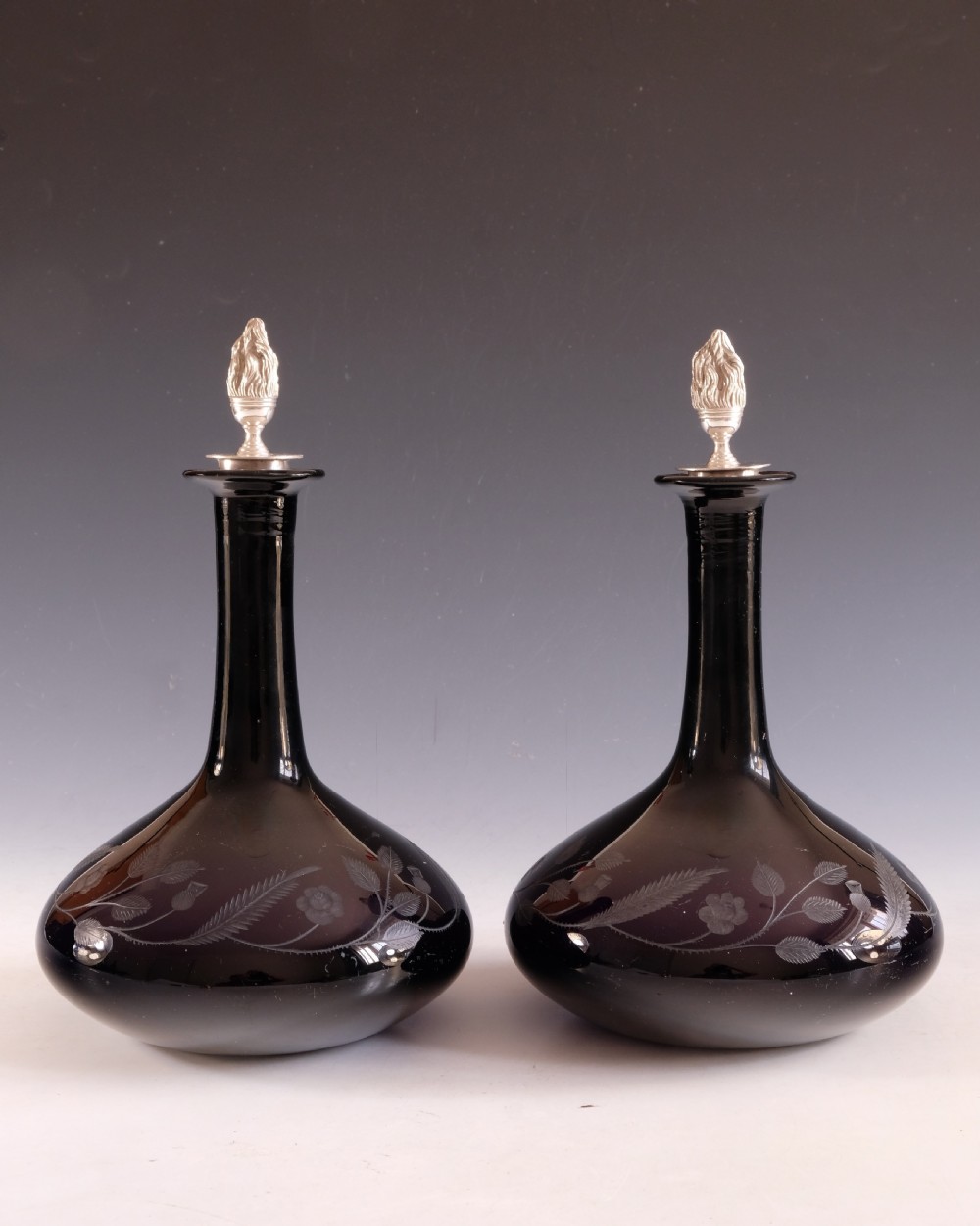 A pair of dark amethyst glass decanters featuring wheel-engraved botanical motifs and topped with silver flame-shaped stoppers. View VIII