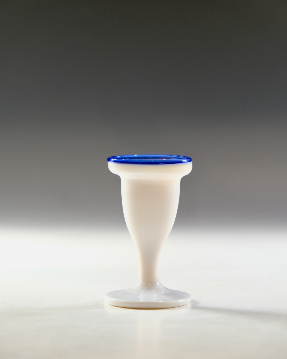 An opaque white glass egg cup featuring a cobalt blue rim on a narrow pedestal stem and flared circular base.