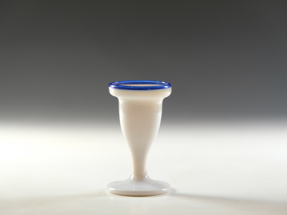 An opaque white glass egg cup featuring a cobalt blue rim on a narrow pedestal stem and flared circular base. View II