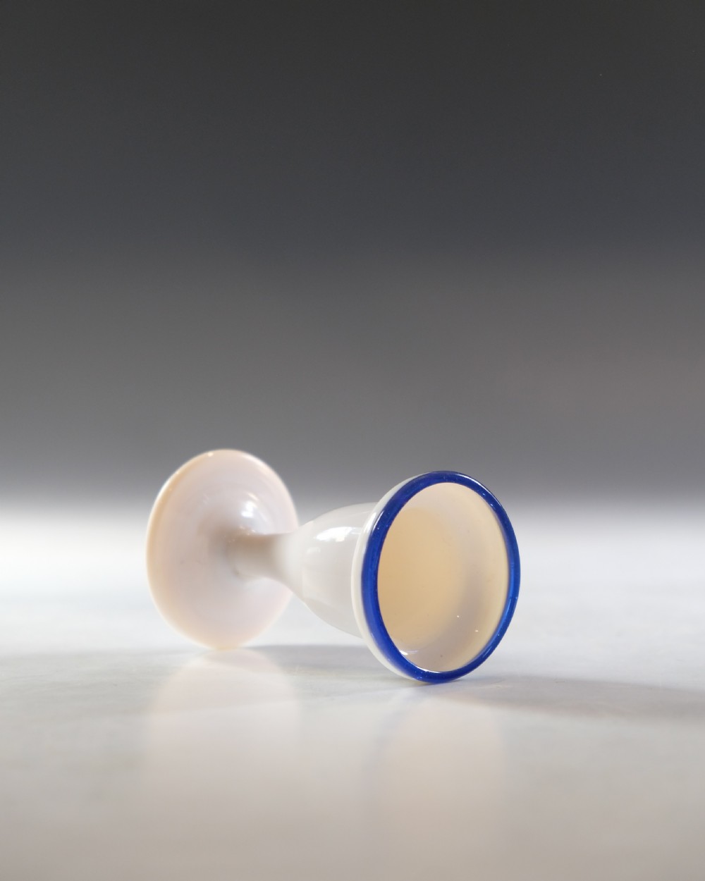 An opaque white glass egg cup featuring a cobalt blue rim on a narrow pedestal stem and flared circular base. View IV
