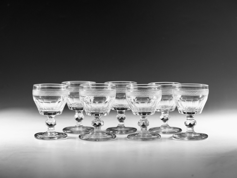 A set of seven clear Georgian cut glass port glasses featuring diamond-patterned bands and faceted stems.