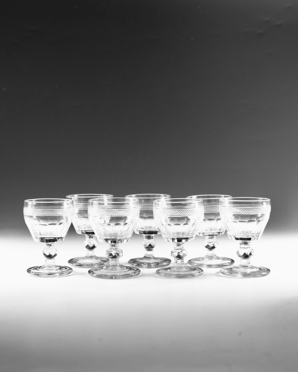 A set of seven clear Georgian cut glass port glasses featuring diamond-patterned bands and faceted stems. View II