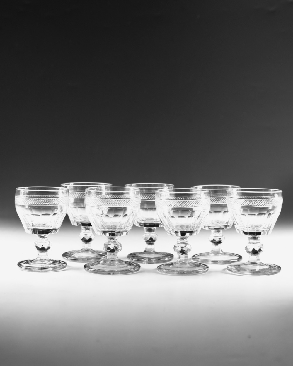 A set of seven clear Georgian cut glass port glasses featuring diamond-patterned bands and faceted stems. View III