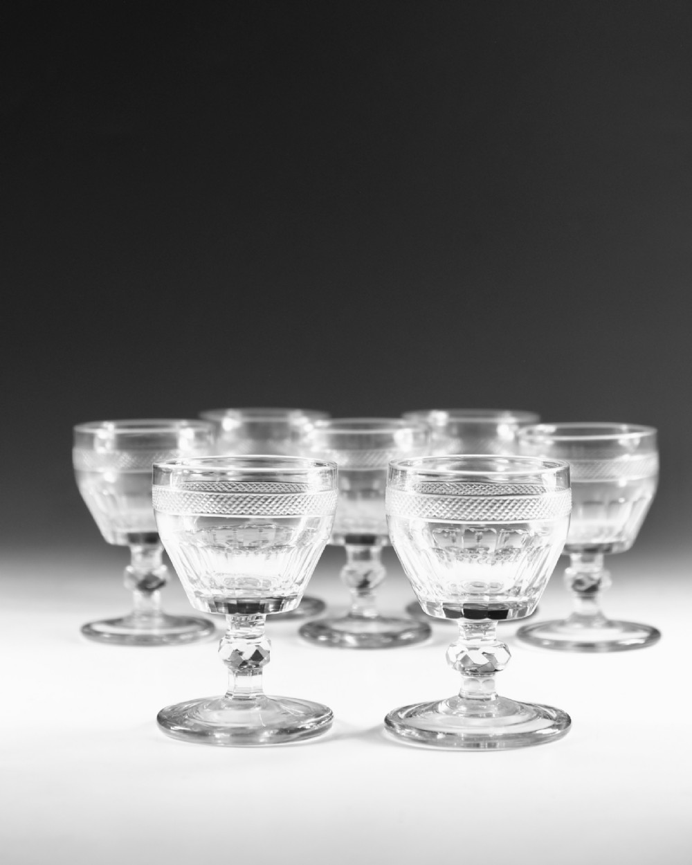 A set of seven clear Georgian cut glass port glasses featuring diamond-patterned bands and faceted stems. View IV