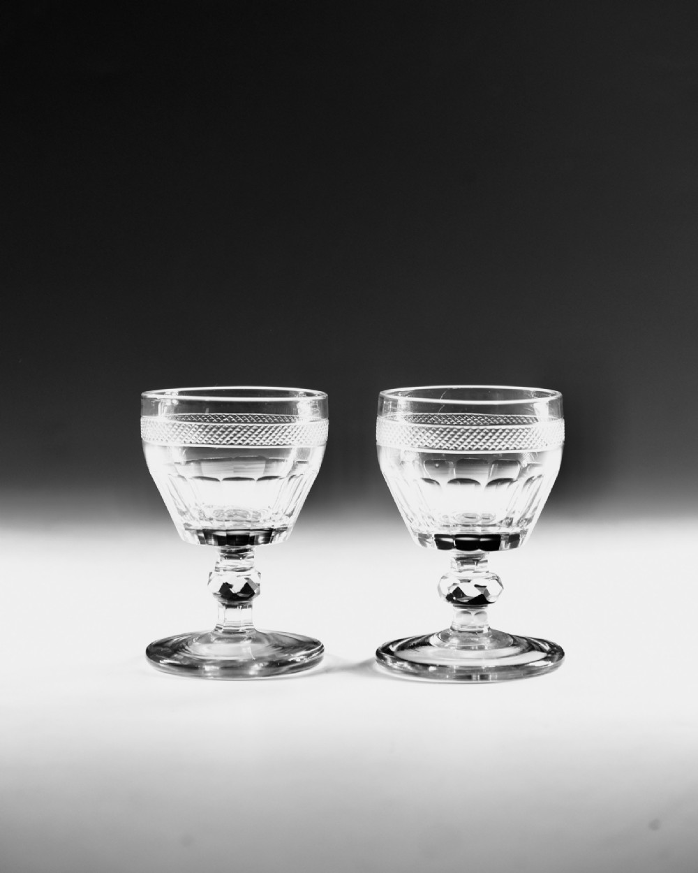 A set of seven clear Georgian cut glass port glasses featuring diamond-patterned bands and faceted stems. View V