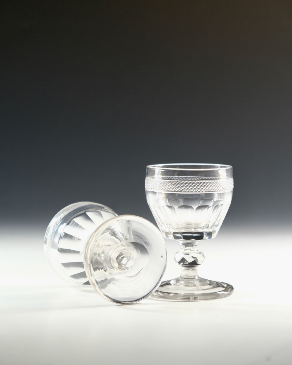 A set of seven clear Georgian cut glass port glasses featuring diamond-patterned bands and faceted stems. View VI