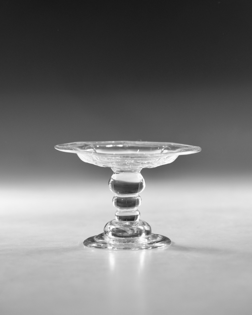 An eighteenth-century Georgian clear lead glass patch stand featuring a shallow rimmed top, a multi-knopped baluster stem, and a circular folded foot. View II