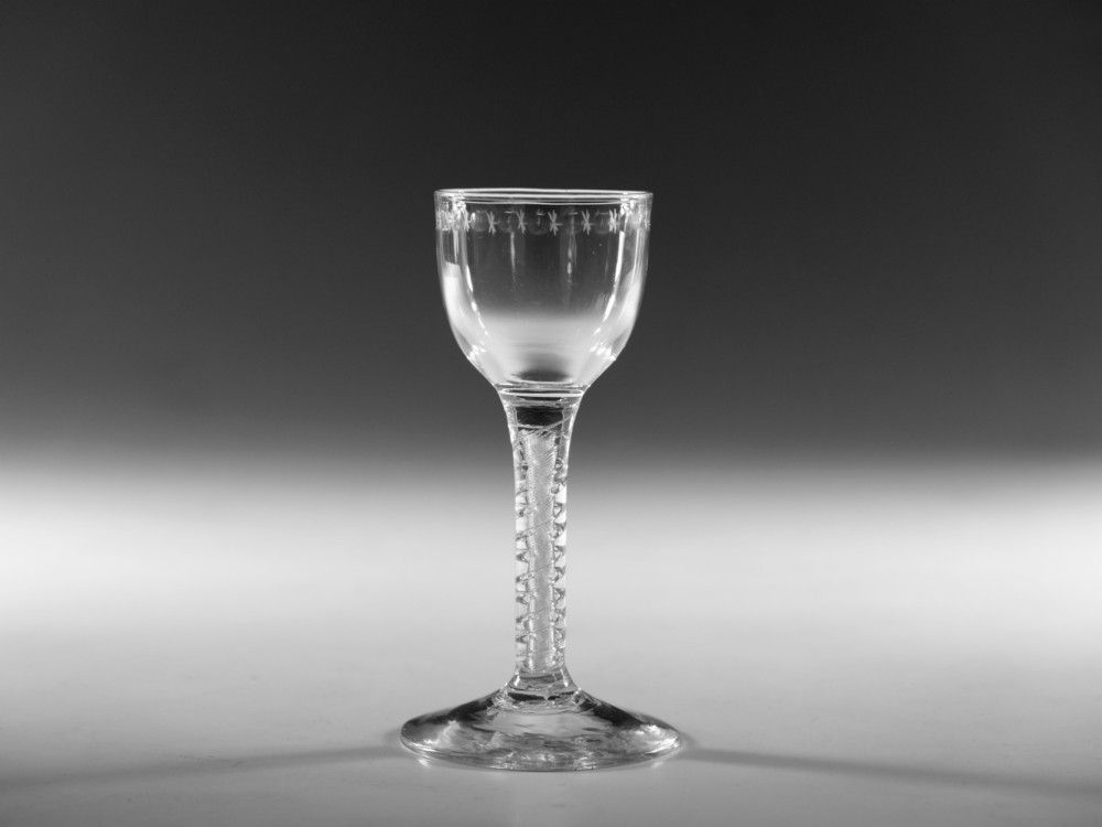 A clear glass wine goblet featuring a wheel-engraved rim pattern, an air twist stem, and a flat conical foot, dating to circa 1755.