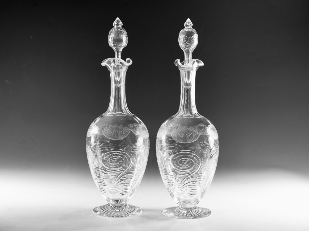 A pair of antique clear glass decanters with intaglio water lily engravings, tall slender necks, and matching glass stoppers.
