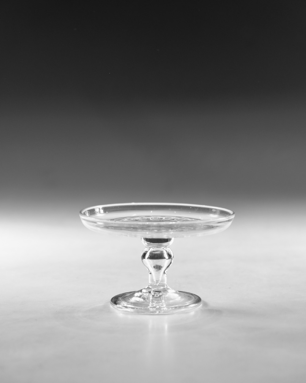 A clear Georgian lead glass patch stand featuring a shallow circular top dish elevated on a baluster stem and a spreading foot. View II