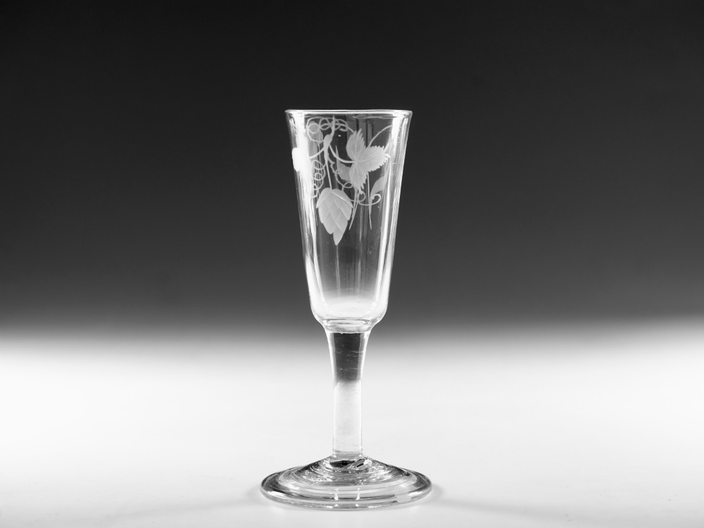 A tall Georgian clear glass ale flute with an engraved hop and barley motif, a straight plain stem, and a flat conical base.