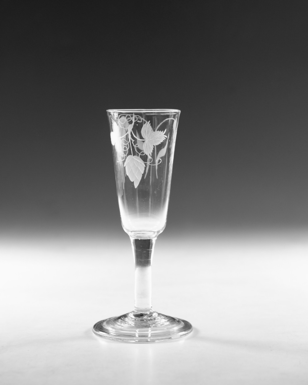 A tall Georgian clear glass ale flute with an engraved hop and barley motif, a straight plain stem, and a flat conical base. View II