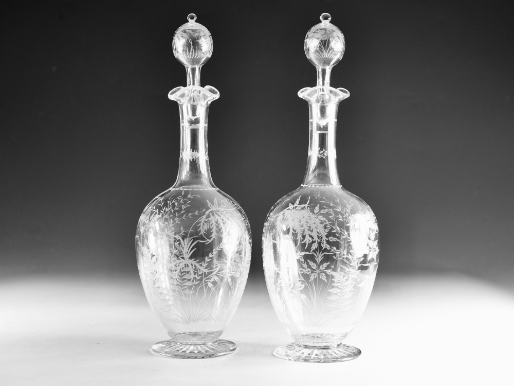 A pair of clear Victorian glass decanters featuring etched botanical fern decoration, narrow necks, ruffled collars, and spherical stoppers.