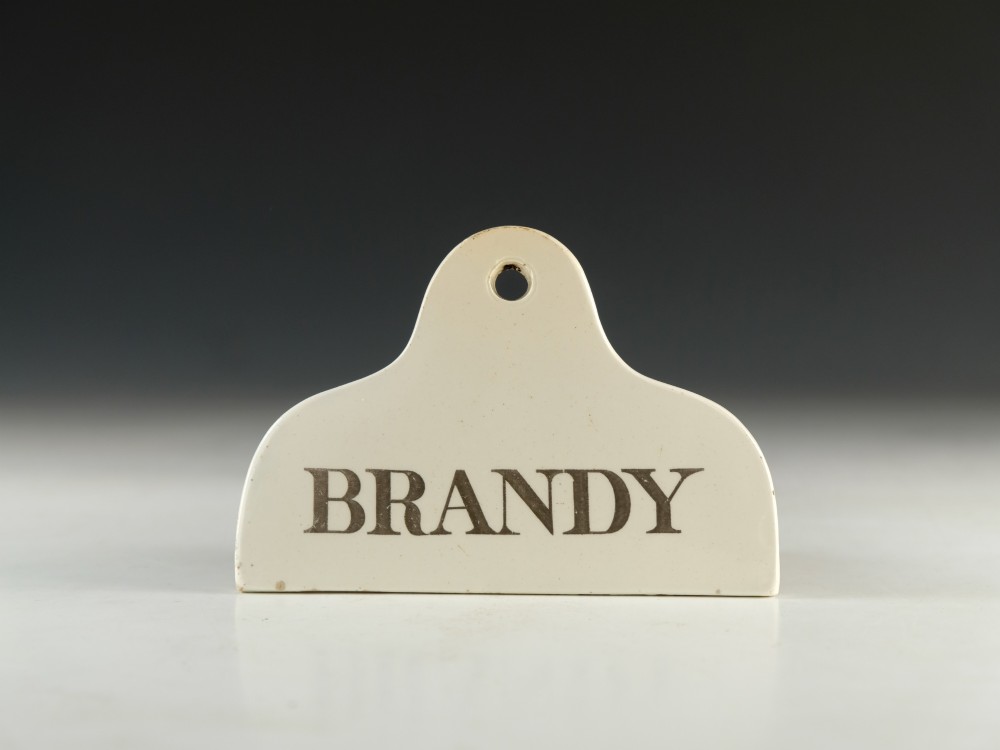 A mid-19th-century cream ceramic brandy bin label featuring black serif lettering and an arched crest with a central hanging hole.