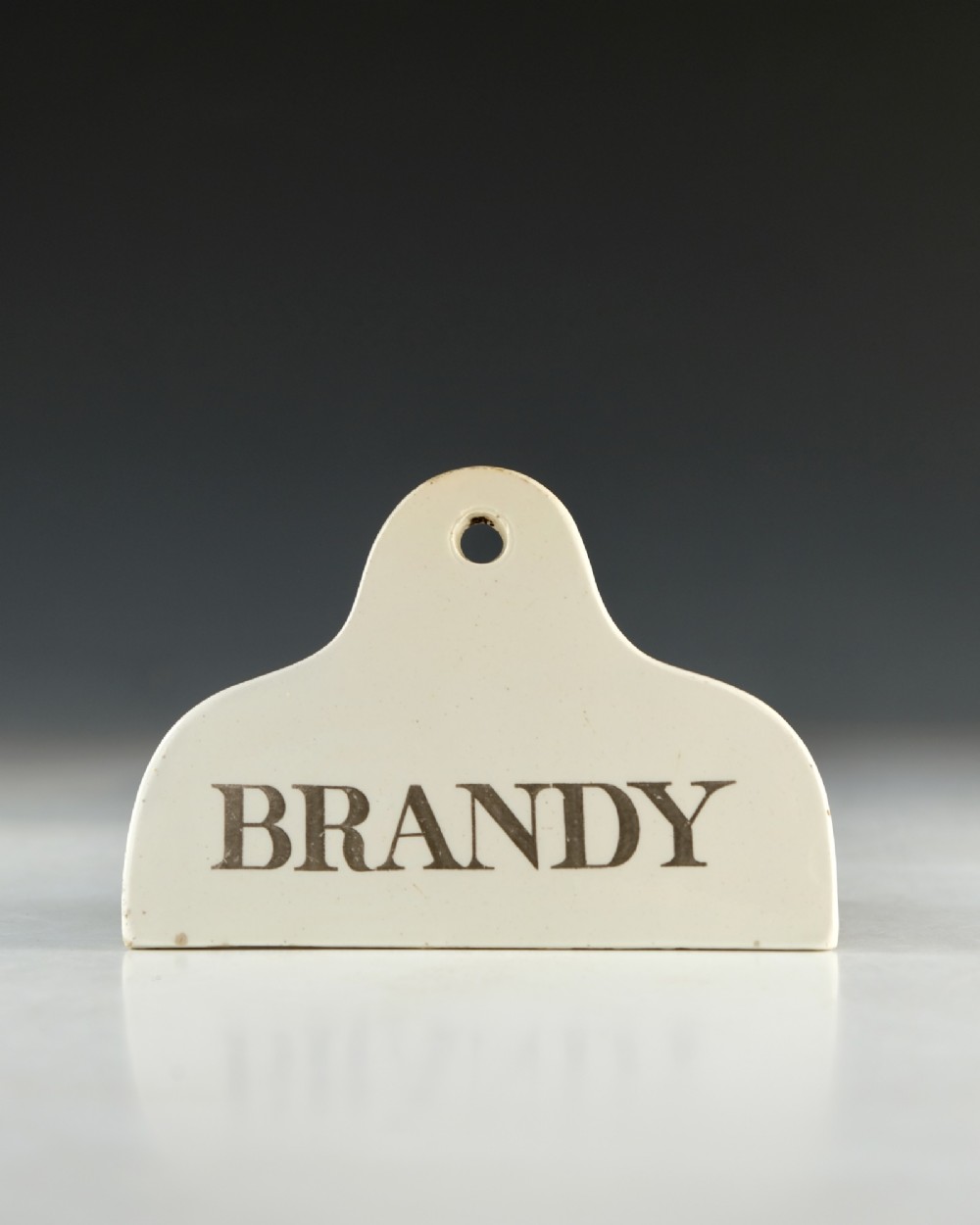 A mid-19th-century cream ceramic brandy bin label featuring black serif lettering and an arched crest with a central hanging hole. View II