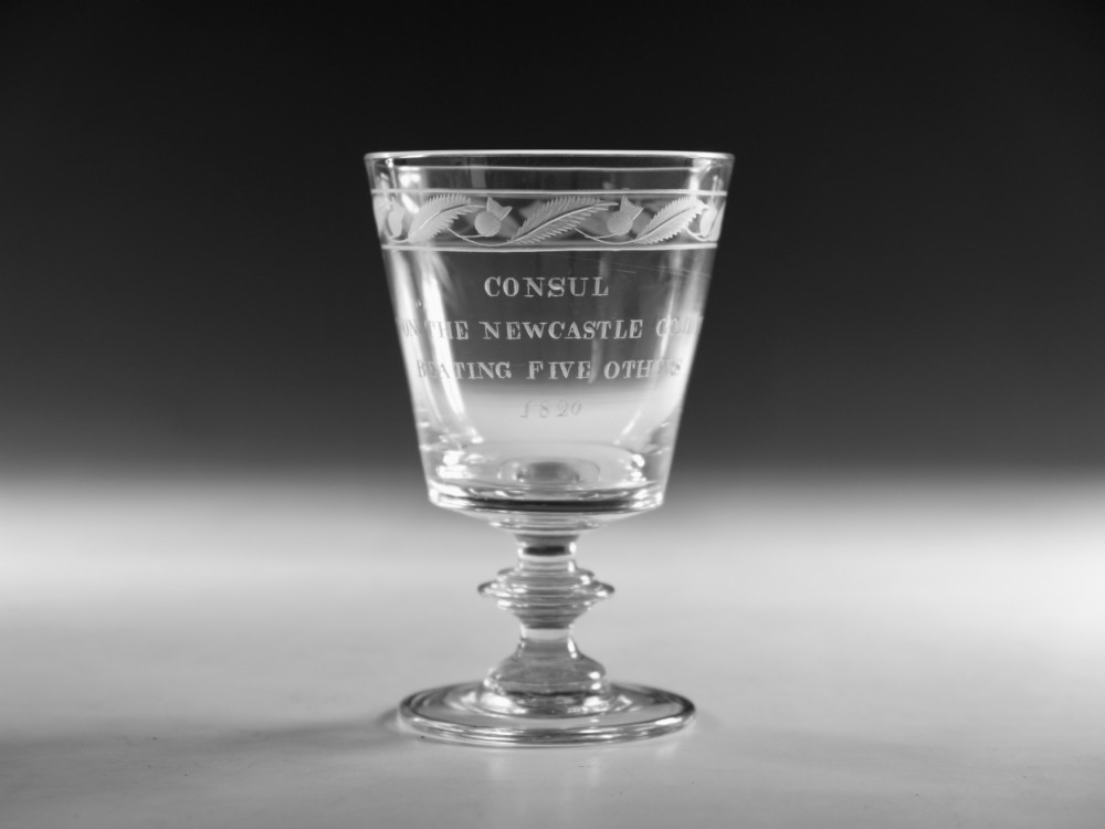 An antique clear glass Georgian rummer featuring wheel-engraved inscriptions and foliage motifs above a knopped stem.