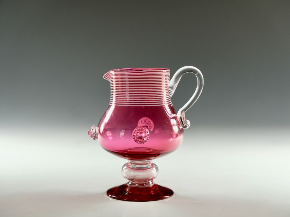 A Victorian cranberry glass cream jug with white threaded details on the neck, a clear applied handle, glass prunt decorations, and a pedestal base.