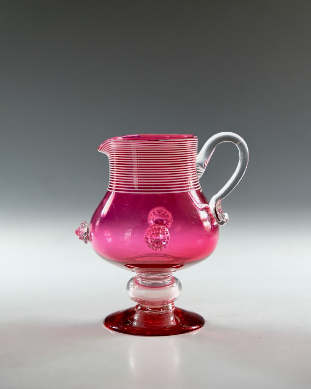 A Victorian cranberry glass cream jug with white threaded details on the neck, a clear applied handle, glass prunt decorations, and a pedestal base. View II