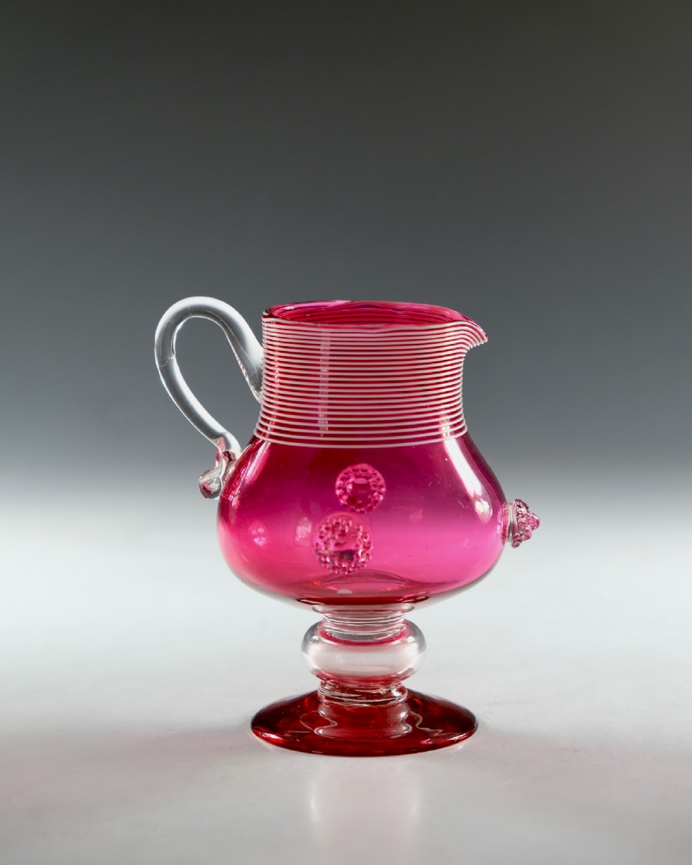 A Victorian cranberry glass cream jug with white threaded details on the neck, a clear applied handle, glass prunt decorations, and a pedestal base. View III