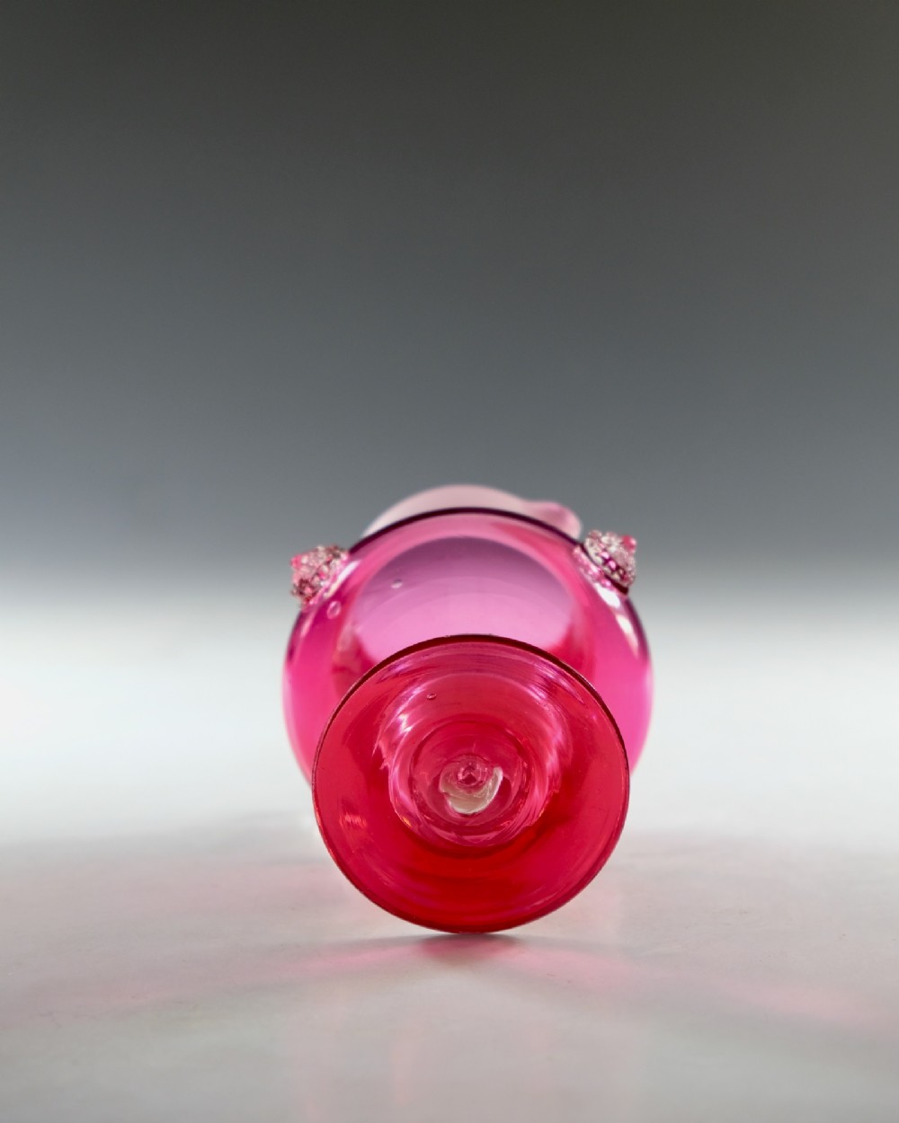 A Victorian cranberry glass cream jug with white threaded details on the neck, a clear applied handle, glass prunt decorations, and a pedestal base. View VI