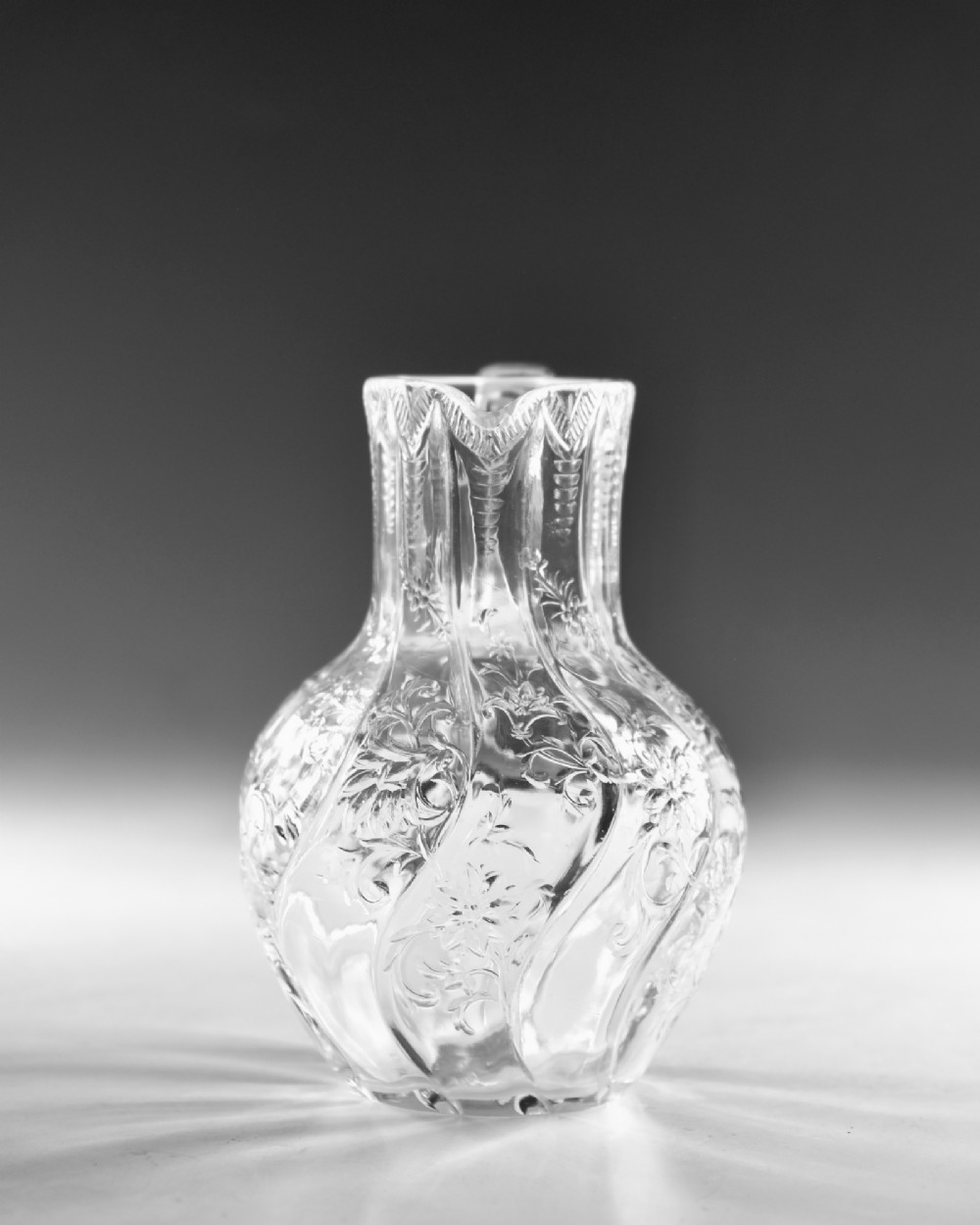 An antique clear glass jug featuring a bulbous swirl-moulded lower body with wheel-engraved floral motifs and a fitted handle. View III