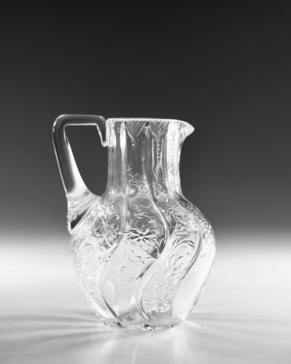 An antique clear glass jug featuring a bulbous swirl-moulded lower body with wheel-engraved floral motifs and a fitted handle. View IV
