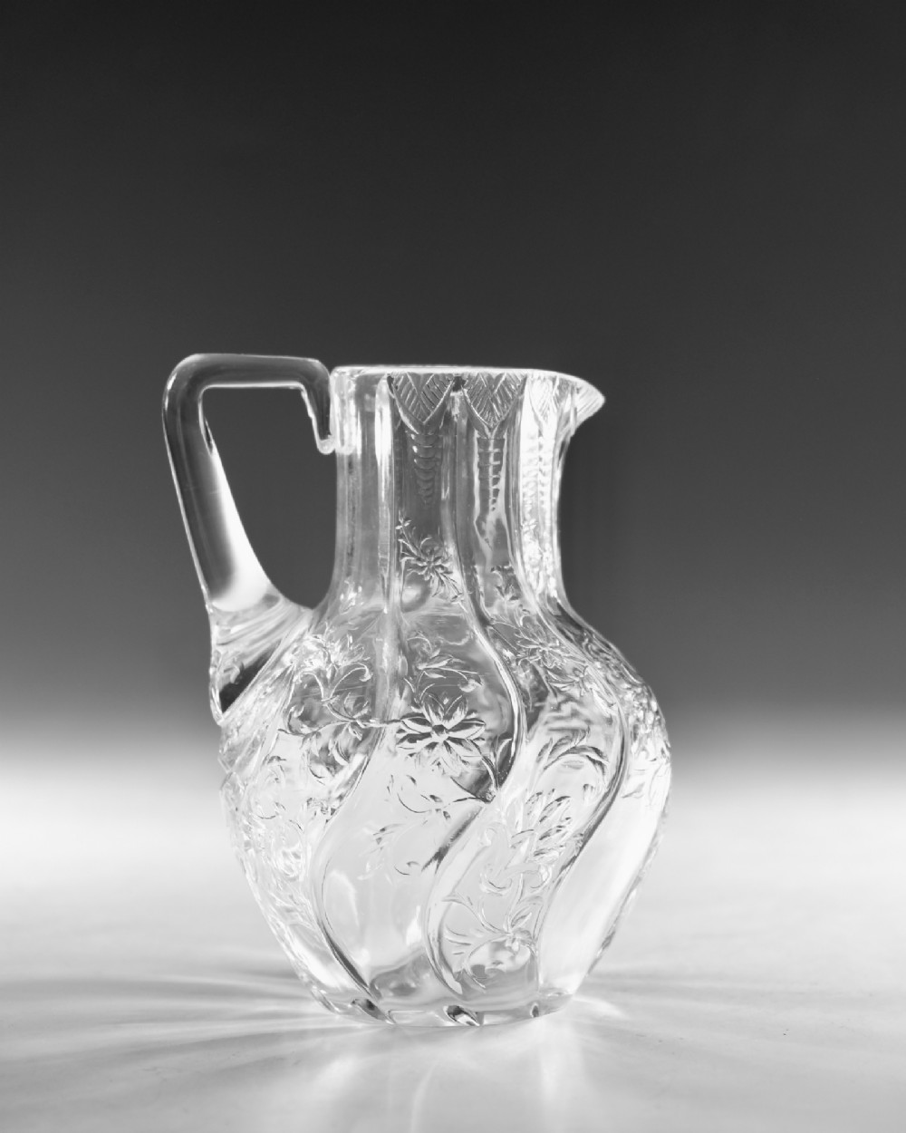 An antique clear glass jug featuring a bulbous swirl-moulded lower body with wheel-engraved floral motifs and a fitted handle. View V