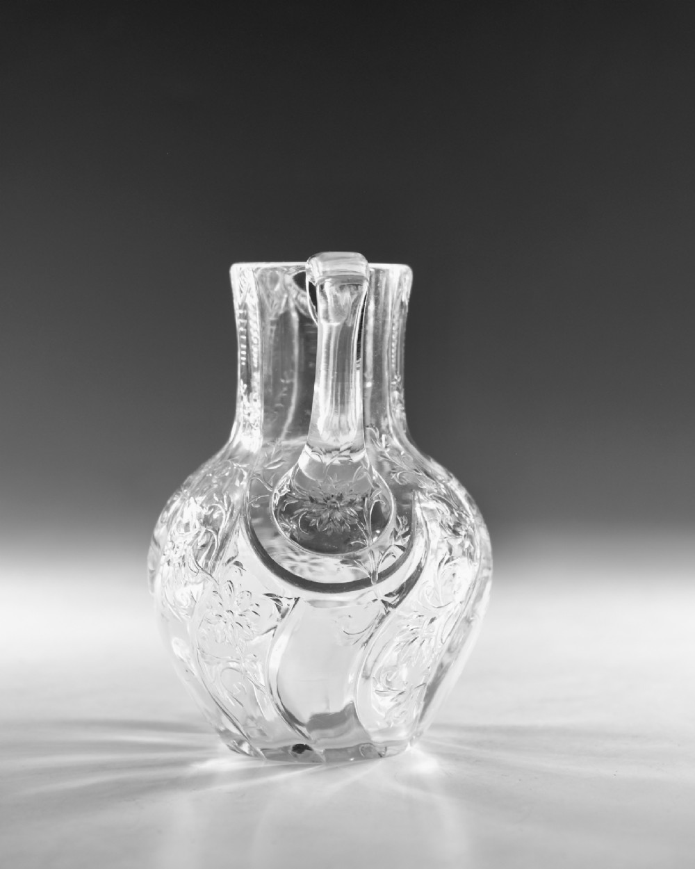 An antique clear glass jug featuring a bulbous swirl-moulded lower body with wheel-engraved floral motifs and a fitted handle. View VI