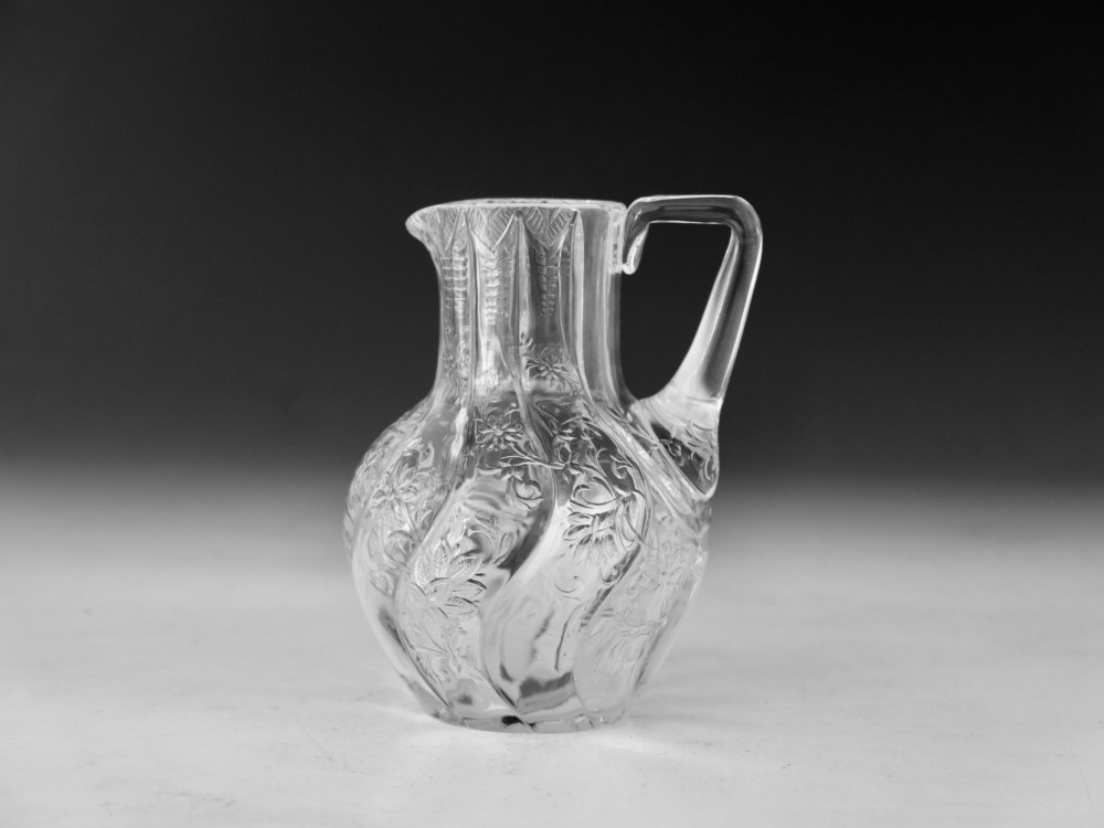 An antique clear glass jug featuring a bulbous swirl-moulded lower body with wheel-engraved floral motifs and a fitted handle. View X