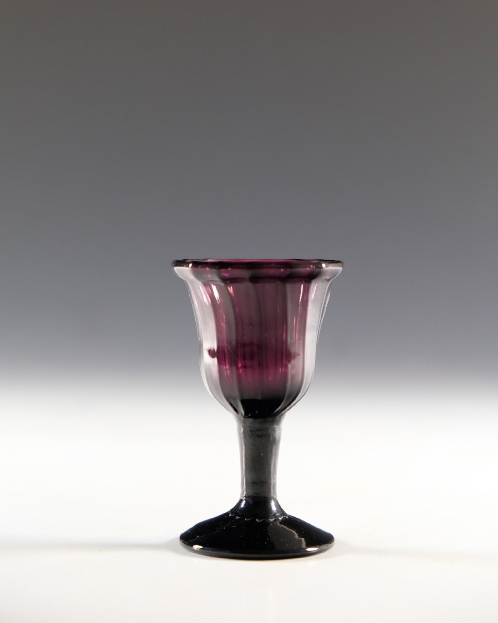 A Georgian amethyst purple glass egg cup with a fluted bowl, slender stem, and circular pedestal foot. View II