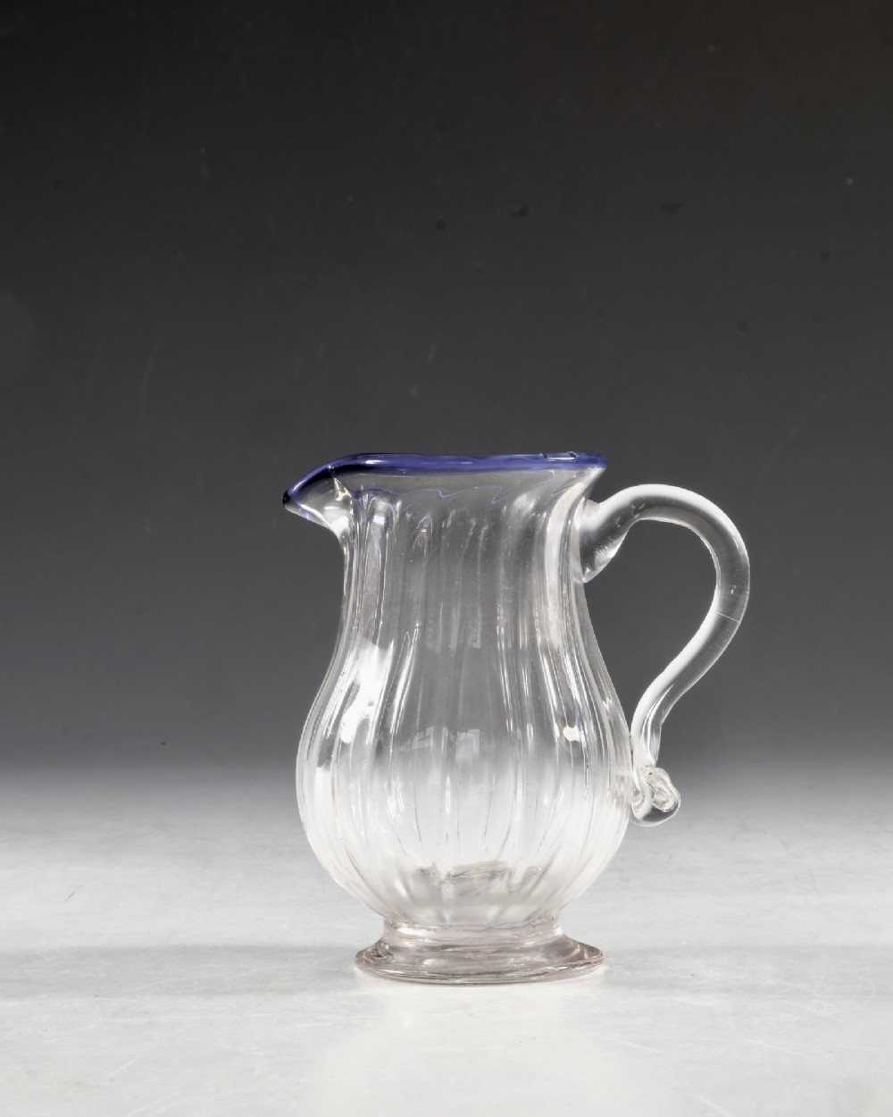 An English Georgian blown clear glass cream jug featuring vertical fluting, a loop handle, and an applied cobalt blue rim. View II